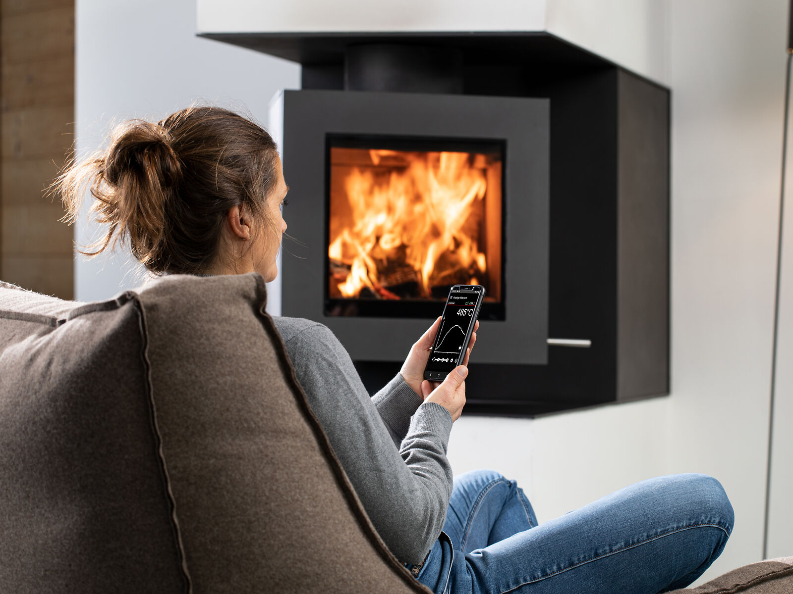 Frau sitzt vor brennendem Drehfeuer. In der Hand ein Smartphone mit geöffneten BRUNNER EAS-App