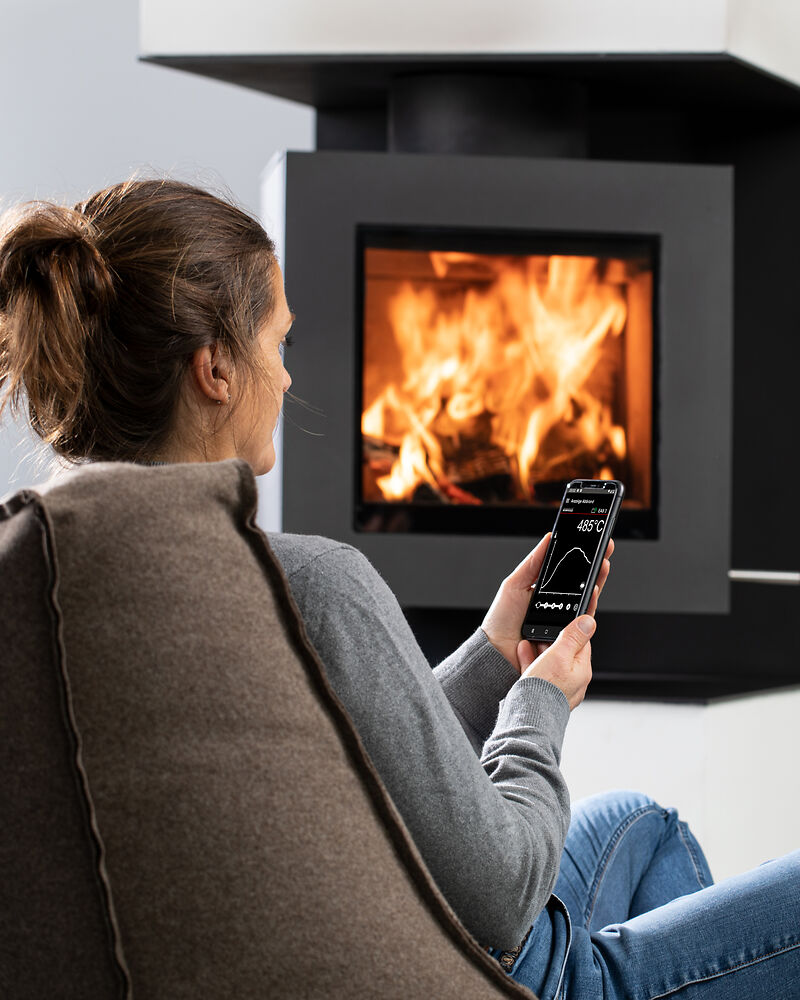 Frau sitzt vor brennendem Drehfeuer. In der Hand ein Smartphone mit geöffneten BRUNNER EAS-App