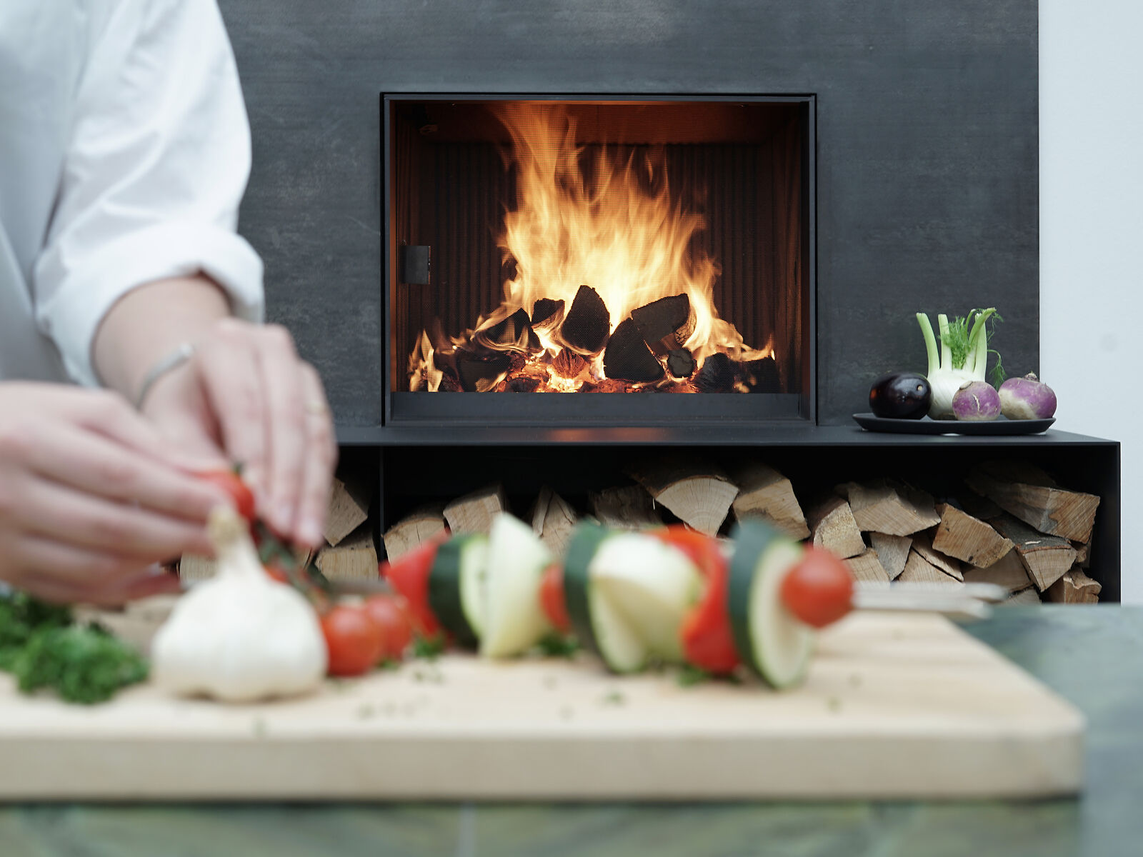 Brennender Urfeuer Küchenkamin mit Person im Vordergrund, die Gemüsespieße vorbereitet