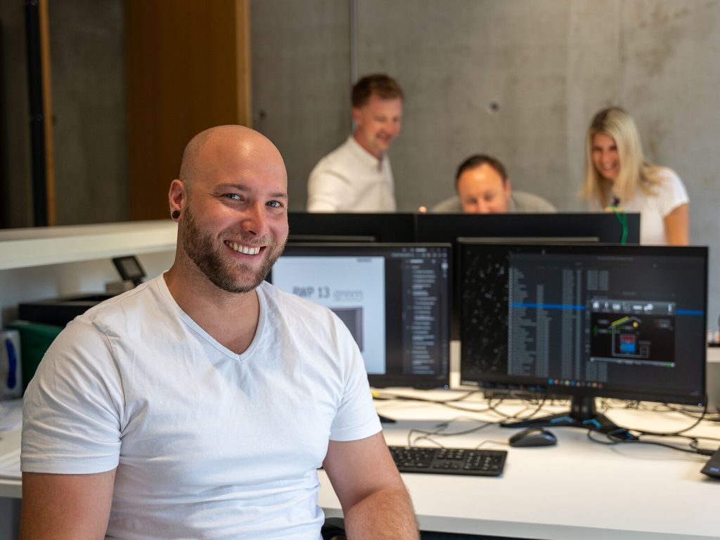 Lächelnder Mitarbeiter sitzt an seinem Schreibtisch. Im Hintergrund besprechen sich zwei Kollegen.