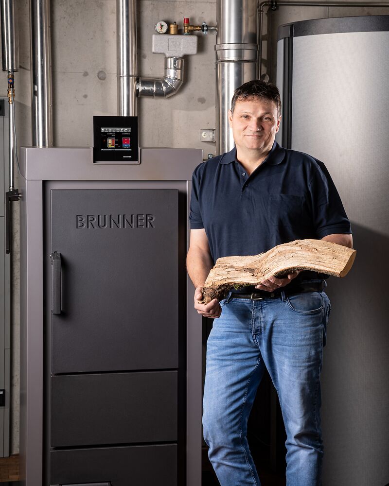BRUNNER Mitarbeiter lehnt am Scheitholzkessel an und hält einen Holzscheit in den Händen