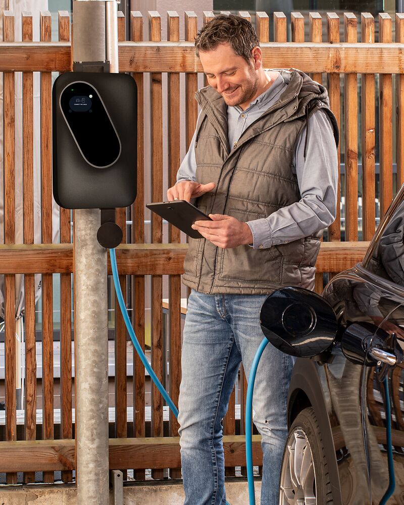 Mann steuert die BRUNNER Wallbox mit einem Tablett und steht neben seinem Auto und der Wallbox